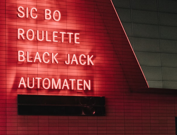 Red neon letters are mounted on a dark tiled wall, displaying the words Poker, Sic Bo, Roulette, Black Jack, and Automaten.