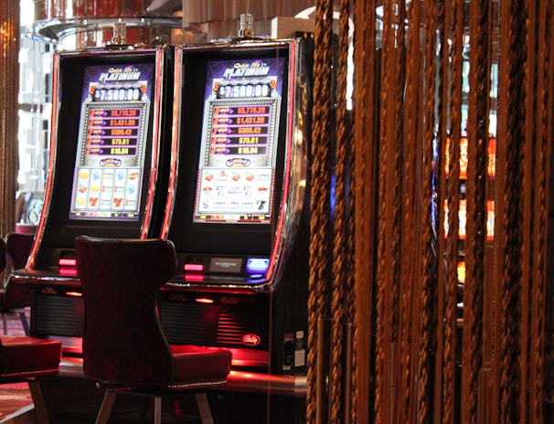 A pair of casino slot machines, each with detailed screens displaying various game outcomes and jackpot amounts. The chairs in front of the slot machines are dark and cushioned for comfort. The background features a hanging curtain made of golden ropes and other casino decorations are visible.
