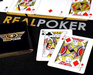 Poker-themed setup featuring a deck of cards on a table with the words 'REAL POKER' visible. Two cards, the King of Clubs and Jack of Diamonds, are prominently displayed. The design is vibrant, with detailed illustrations on the cards.