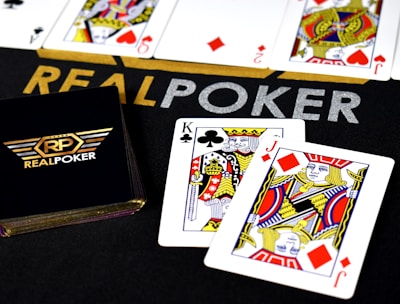 Poker-themed setup featuring a deck of cards on a table with the words 'REAL POKER' visible. Two cards, the King of Clubs and Jack of Diamonds, are prominently displayed. The design is vibrant, with detailed illustrations on the cards.