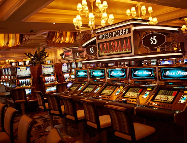 A casino floor featuring rows of brightly lit video poker machines. The area is illuminated by elegant chandeliers, with a display board showing a large sum of money, suggesting a possible jackpot. The rich decor includes luxurious carpets and comfortable seating, adding to the opulent atmosphere.
