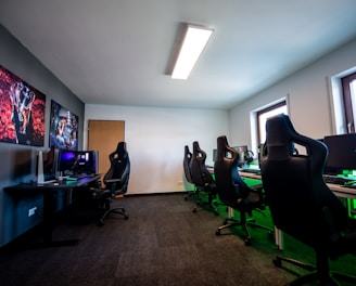 A well-lit gaming room features multiple gaming stations with high-backed chairs and monitors. The room has a minimalist setup with green and black color tones. Large posters adorn the walls, and the room appears clean and organized.