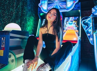 A person sitting on an arcade game machine platform with neon lights in the background. The arcade game is called 'Lane Master' and features a colorful design with bowling-themed imagery.