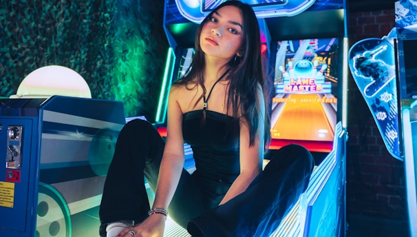 A person sitting on an arcade game machine platform with neon lights in the background. The arcade game is called 'Lane Master' and features a colorful design with bowling-themed imagery.