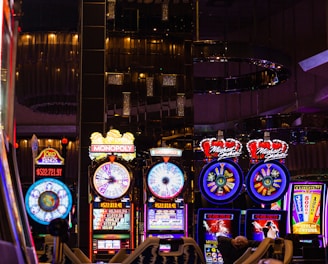 A lively casino scene featuring a row of bright, colorful slot machines with themes like Monopoly, Michael Jackson, and Lord of the Rings. The gaming machines display a variety of flashing lights and digital screens, creating an energetic and exciting atmosphere. Reflections of the lights are visible in glossy surfaces above the machines.