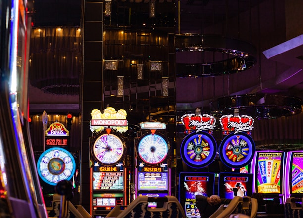 A lively casino scene featuring a row of bright, colorful slot machines with themes like Monopoly, Michael Jackson, and Lord of the Rings. The gaming machines display a variety of flashing lights and digital screens, creating an energetic and exciting atmosphere. Reflections of the lights are visible in glossy surfaces above the machines.
