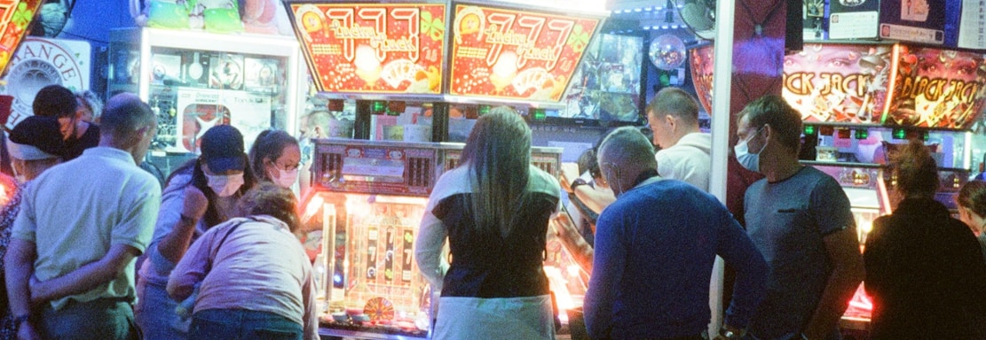 A group of people standing in front of an illuminated amusement arcade, engaging with various gaming machines. The environment is bustling with activity, and colorful lights create a vibrant atmosphere. The ceiling is decorated with large, playful images of dice and other game-related graphics, enhancing the lively mood.