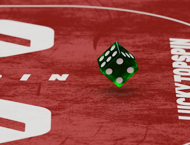 Two green dice with rounded edges are positioned on a red surface that seems to be part of a game board. The surface has white text and numbers on it, including the word 'LUCKYTOPSPIN' in capital letters.