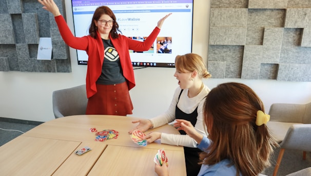 Four people are engaged in a lively game around a wooden table. One person stands with raised arms, while three others are seated, focusing on colorful cards in their hands. There is a computer monitor in the background displaying a webpage, and the room features a modern design with gray acoustic panels on the walls.